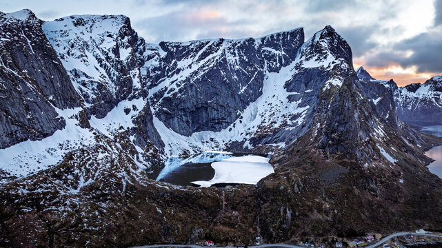 Aerial view of majestic snow-capped mountains pierce the sky, their peaks softened by the pastel dawn light, casting long shadows over the still waters below, Reine, Nordland, Norway.
