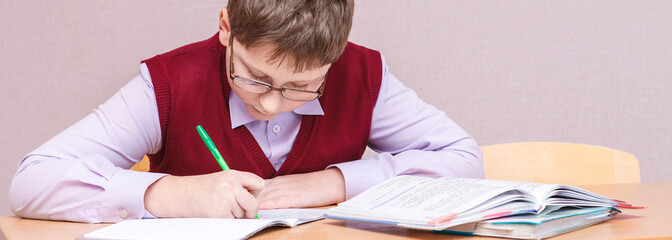 boy with glasses sitting at a desk writing. in glasses and school uniform