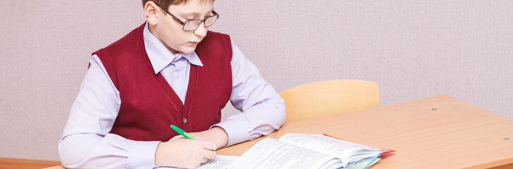 boy with glasses sitting at a desk writing. in glasses and school uniform