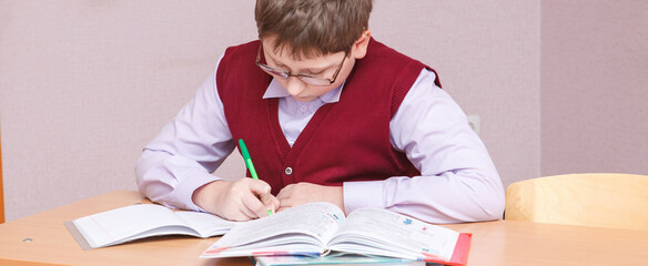 boy with glasses sitting at a desk writing. in glasses and school uniform