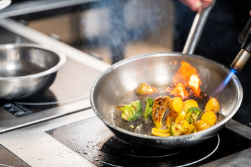 Chef searing Brussels sprouts and potatoes with a blowtorch in a stainless steel pan on an induction cooktop, showcasing dynamic cooking action in a professional kitchen.