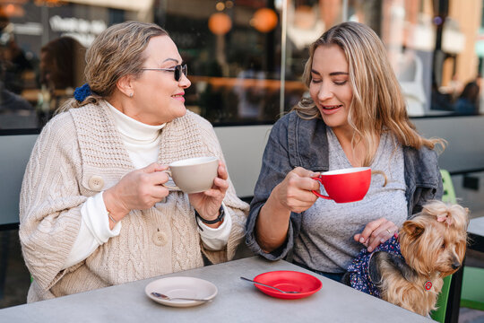 Two women enjoy coffee at a bustling outdoor cafe while a small dog sits in a cozy atmosphere on a sunny day in the city - Powered by Adobe