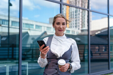 Businesswoman in a stylish outfit checking her smartphone while holding a coffee cup outside a...