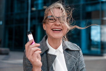 Woman in stylish grey coat laughing outdoors while holding a vape device near modern building on a...