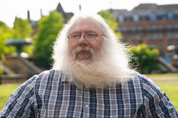 Elderly man with long white beard smiles in a park with greenery and buildings in the background during a sunny day