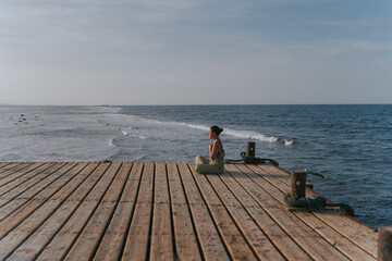 Young woman meditating on a wooden pier at sunset by the sea