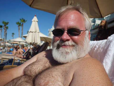 Relaxing older chubby man with beard enjoying sunny day at hotel poolside, surrounded by palm trees and umbrellas, creating cheerful holiday