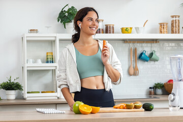 Athletic woman eating carrot and preparing smoothie while listening to music with earphones in the...