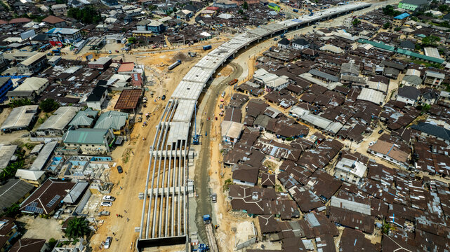 Aerial view of a gray concrete construction site cuts through a dense urban landscape, juxtaposing new infrastructure with the existing residential buildings, Lagos, Nigeria.