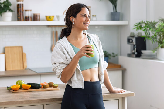 Kind sporty woman drinking a healthy green smoothie standing in the kitchen at home.