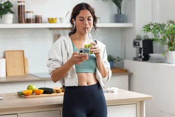 Sporty woman drinking a healthy green smoothie while using smartphone in the kitchen at home.