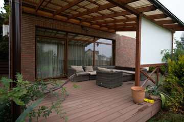 Fototapeta premium Cozy Wooden Patio with Rattan Furniture and Greenery Around a Modern Brick Home