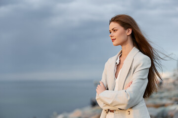 Woman in beige coat standing confidently outdoors with arms crossed, natural makeup, long hair...
