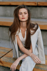 woman with long brown hair in white suit sitting on wooden stairs confident serious look fashion...