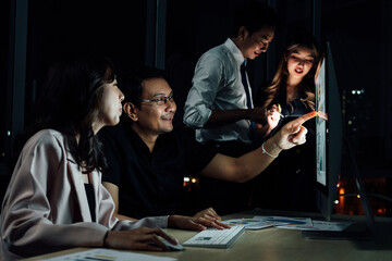 Group of Southeast Asian professionals analyzing data on desktop monitor during nighttime in corporate office, showing determination and focus under dim ambient lighting.
