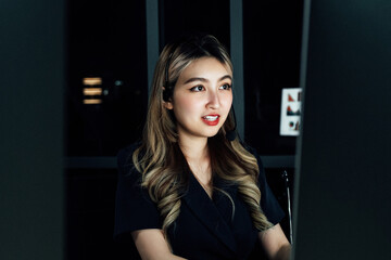 Asian female call center agent working night shift in dimly lit office, wearing headset and...