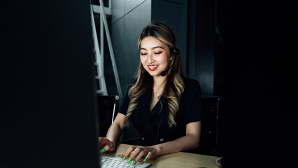 Fototapeta premium Professional Asian woman in headset assisting client during night shift, illuminated by computer light in dark workspace, representing 24/7 customer support and communication skills. 
