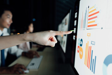 Close-up of female hand pointing at screen with analytics, colleagues working on computers in background. Business tech concept.