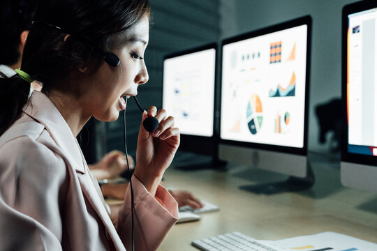 Asian female call center agent with headset and microphone smiling while assisting customer, with blurred colleagues in background. Concept of customer support and workplace technology.