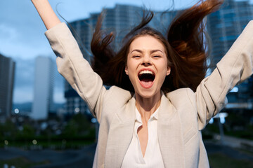 Excited woman in beige jacket celebrating success in urban business district with joyful expression...
