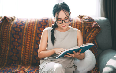 Beautiful young adult asian woman wearing eyeglasses reading book on sofa relax hobby routines