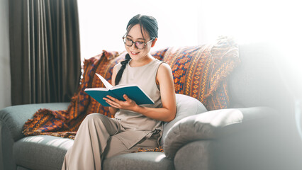 Beautiful young adult asian woman wearing eyeglasses reading book on sofa relax hobby routines