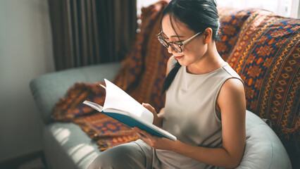 Beautiful young adult asian woman wearing eyeglasses reading book on sofa relax hobby routines