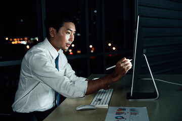 Focused Asian businessman working late at night, analyzing data on digital tablet screen. Business report with colorful graphs displayed on monitor.

