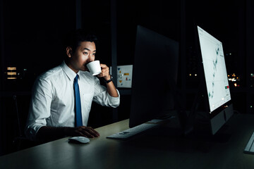 Asian businessman staying up late while drinking coffee and analyzing business charts on desktop screen. Night office illuminated only by monitor.