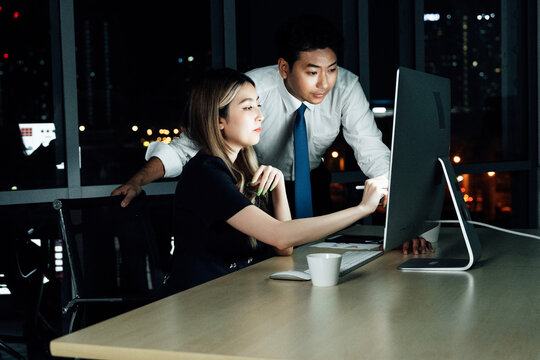 Asian business colleagues working late night at office, reviewing project details on desktop screen under city lights, showing dedication and teamwork.