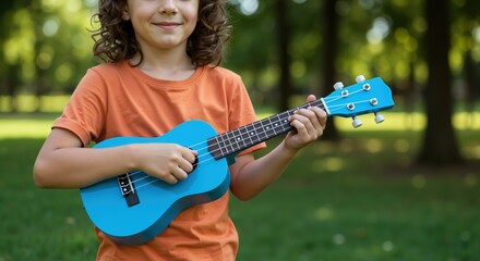 Girl with curly hair playing blue ukulele in orange shirt outdoors. Child learning musical instrument in green park setting. Music education and outdoor learning concept
