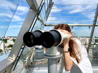 Infront view of a teenage girl looking through stationary binoculars at cityscape from observation...