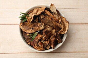 Pieces of dried mushrooms and rosemary in bowl on light wooden table, top view