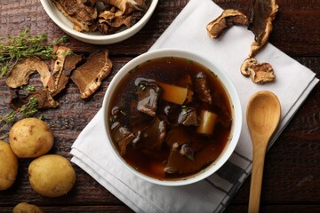 Tasty soup in bowl, raw potato and dry chanterelle mushrooms on wooden table, flat lay