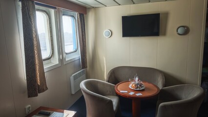 Cozy ferry cabin interior with two windows, round table, armchairs and a TV on the wall, daylight...