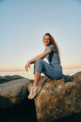 Lifestyle woman in modern boho-western denim street style sitting on rocks by the sea during golden hour with film color warmth