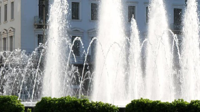 Dynamic water jets shoot upward from a city fountain in Omonia Square, Athens, Greece. Sunlight reflects off the spray. Light-colored building and green foliage form the background. No people visible