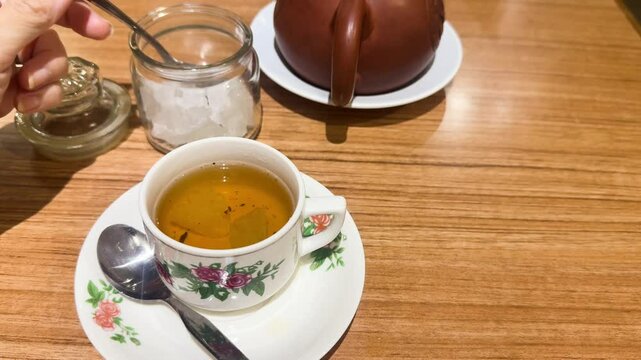 Teh Poci, traditional Javanese jasmine tea from Central Java, served in a floral porcelain cup with rock sugar and clay teapot. A spoon adds sugar in this cozy Indonesian tea ritual.