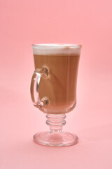 Elegant glass mug filled with creamy coffee against a pastel pink background