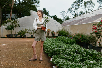 Young female gardener in casual outfit smiling with potted plant in greenhouse, exuding joy in...