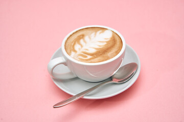 Latte art in white cup on saucer with spoon against pink background