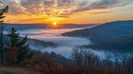 Fototapeta premium Sunrise over misty forested hills, with golden light breaking through a partly cloudy sky and fog filling the valley below.