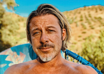 Happy tanned blond man with grey stubble beard sits in a blue chair outside in hilly and sunny...