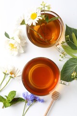 Aromatic herbal tea in glass cups, different flowers and green leaves on white background, flat lay