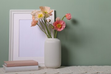 Beautiful poppy flowers in vase, books and empty frame on table near green wall, closeup