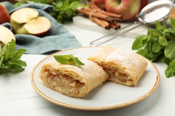 Pieces of tasty apple strudel with powdered sugar, mint, fruits and cinnamon on white marble table,...
