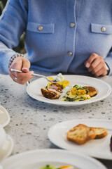 Person eating breaded meat with potatoes and creamy sauce at a dining table, surrounded by fresh bread and vegetables. Close-up view of casual meal time.