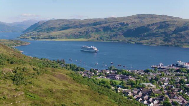 Drone shot of Viking Sun cruise ship anchored in the loch near Ullapool, with stunning views of the village, surrounding mountains, and clear blue skies on a beautiful day.