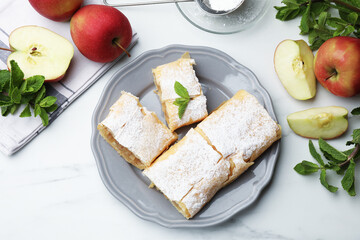 Pieces of tasty apple strudel with powdered sugar, mint and fruits on white marble table, flat lay