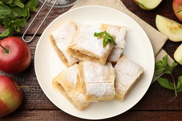 Pieces of tasty apple strudel with powdered sugar, mint and fruits on wooden table, flat lay